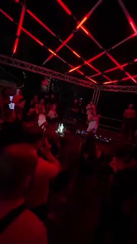 A crowd of people are gathered in a dimly lit outdoor venue, illuminated by red neon lights arranged in a geometric pattern on the ceiling. A DJ is visible on a raised platform in the background, with speakers and equipment set up. Some individuals in the crowd are dancing and interacting with each other, while others are standing and observing. The atmosphere appears to be that of a live music event or party.