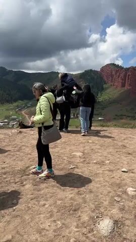 The video shows an outdoor scene in Jeti-Ögüz, Kyrgyzstan, featuring several people and a horse in a mountainous landscape.

In the center, a woman with blonde curly hair, wearing a blue jacket, grey pants, and sunglasses, is seated on a black horse equipped with a patterned saddle. She is smiling and appears to be adjusting her position. A man in a black long-sleeved shirt, black pants, and a grey sling bag with a "depe" branded water bottle attached, stands next to the horse, assisting her. To the right of the horse, another woman in a black jacket and light blue jeans is standing, observing. In the foreground, a woman in a light green jacket, black pants, and a grey shoulder bag walks across the frame.

The group is on a dusty, brown hilltop or plateau. The background features rolling green hills and mountains covered in trees. Distinctive red rock formations, characteristic of Jeti-Ögüz, are visible on the far right. In the valley below, a small village or settlement with houses can be seen. The sky is partially cloudy, suggesting a variable weather day. The overall impression is of a group engaging in horseback riding amidst picturesque natural scenery.