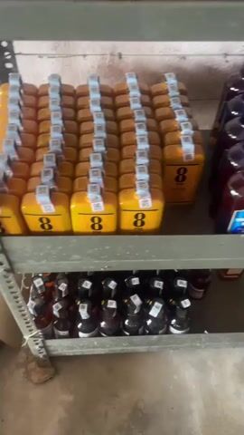 The video shows several shelves of bottled alcoholic beverages. The shelves are made of metal and appear to be in a storage area. On the top shelf, there are rows of yellow bottles with the number 8 on them and several bottles with blue caps that are unclear. On the middle shelf, there are rows of bottles with blue caps and labels that say "Imperial Blue". There are also some brown boxes and bottles with green labels. The bottom shelf contains more bottles with dark liquids and black caps. A broom is visible on the floor in the lower left corner. The lighting is dim, suggesting an indoor environment. There are no people visible in the video.