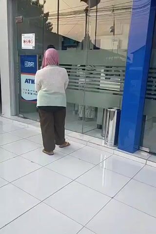 A person wearing a pink hijab and a light-colored cardigan is standing outside a bank. The bank has glass doors with blue accents. There is a sign on the door that says "TUTUP", which means "CLOSED" in Indonesian. There is also an ATM machine visible on the left side of the entrance. A metal trash can is located next to the entrance. The person is facing away from the camera, looking towards the bank entrance. The ground is tiled with white square tiles.