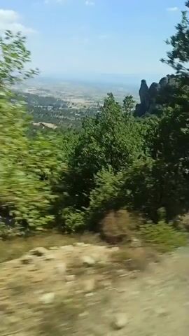The video shows a moving shot from inside a vehicle, looking out at a landscape. The foreground is a dirt road with dry grass and some rocks. Beyond that, there is a dense green forest covering a hillside. In the distance, a valley unfolds with a town or village nestled among more trees and fields. Two prominent rock formations stand tall on the right side of the valley. The sky is blue with a few white clouds. The camera motion suggests the vehicle is traveling along a winding road with an upward incline.
