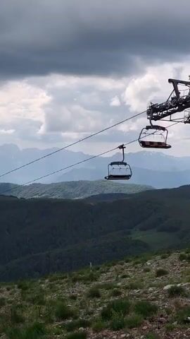 A ski lift ascends a mountain on a cloudy day, with a line of empty chairlifts moving across the frame. Below the ski lift, a lush green forest covers the mountain slopes. In the distance, layers of mountains fade into the cloudy sky. The ground in the foreground is covered in green grass and some sparse rocks.