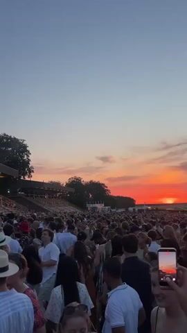 The video shows a large crowd of people gathered outdoors for a concert. The sun is setting, casting a warm glow over the scene. In the background, a large stage is set up with lighting and speakers, and performers can be seen on stage. The crowd is facing the stage, with many people holding up their phones to record or take pictures. Some people are dancing, while others are standing and watching the performance. The overall atmosphere appears to be festive and energetic.