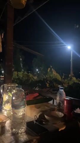The video opens with a close-up of a table at night. A pitcher with a floral design, a plastic water bottle with a blue label, and a red can are visible. In the background, a streetlamp illuminates a patch of greenery and a dark sky. The camera pans slightly, revealing more of the surrounding foliage, a dark road, and what appears to be some construction equipment or debris. The scene is dimly lit, with the streetlamp providing the primary light source. The overall impression is that of an outdoor setting at night, possibly a restaurant or a backyard area adjacent to a rural or semi-rural road.