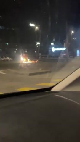 The video shows a scene at night, likely from inside a car. A Renault dealership is visible across the street. In the middle of the road, there appears to be construction work or some other activity that is producing sparks, potentially from grinding or welding. There is at least one worker visible in a yellow vest. Other cars are parked nearby and streetlights illuminate the scene. The road appears wet, perhaps from rain. The car the video is taken from begins to accelerate and drive away.