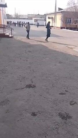 The video shows a schoolyard during the day. A few people are walking around, some children are playing in the background. The ground is cracked asphalt with patches of what might be oil stains. Buildings surround the yard. The lighting suggests a sunny day.