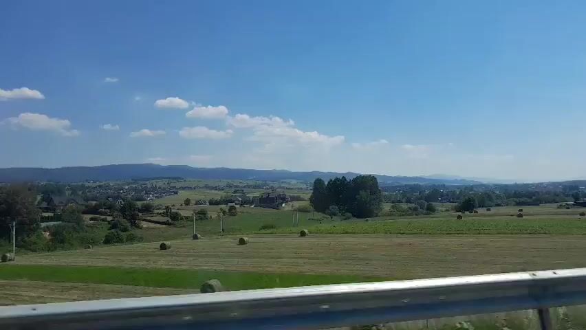 The video shows a view from a vehicle moving through a rural landscape on a sunny day with a clear blue sky and scattered white clouds. The landscape consists of rolling hills, fields of hay bales, trees, and a small village in the distance. The vehicle appears to be on a road with a metal guardrail in the foreground. A sound barrier is visible on the right side of the frame.