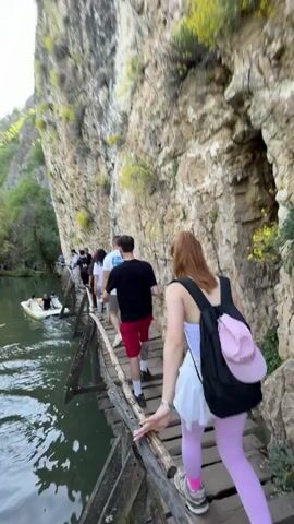 A group of people walks along a narrow wooden bridge built along a cliff face. The bridge is above a body of water, and a small boat with people in it is visible. The group consists of several people, including a woman in the foreground with red hair, a white skirt, and pink leggings. She carries a black backpack with a pink cap attached. The bridge looks old and is made of wooden planks. The cliff face is tall and rocky, with some greenery growing on it. The water is calm with some ripples. The sky appears bright, indicating daylight. The video is shot from slightly behind the walking group.