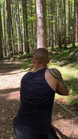 A man with tattoos on his arm is standing in a forest and aiming a rifle. He is wearing a dark blue tank top and black shorts with a white stripe. The ground is covered in dirt and fallen leaves. There are many tall trees around him, and sunlight is filtering through the leaves.