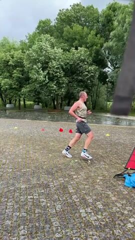 The video depicts a man running an obstacle course outside, likely in a park or similar paved area. The man is wearing shorts and a tank top. He runs over a red ladder-like obstacle, weaves around small cones, and continues running. A second man is filming him with a smartphone, following him as he runs. Other people are visible standing near a table with supplies, possibly water. The weather appears to be overcast and wet, as the ground is damp and shiny.  Trees surround the paved area.