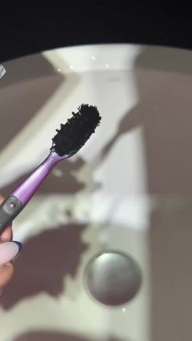 In a close-up, top-down shot, a person with manicured nails, painted with light pink and navy blue tips, holds a purple and gray toothbrush in their left hand. The toothbrush has a black head. With their right hand, the person squeezes a silver tube of toothpaste onto the toothbrush bristles. The toothpaste is white. The scene is set against a white porcelain sink with a shiny silver drain. The lighting casts shadows, creating a slightly surreal or stylized effect. The overall focus is on the act of applying toothpaste to the toothbrush.