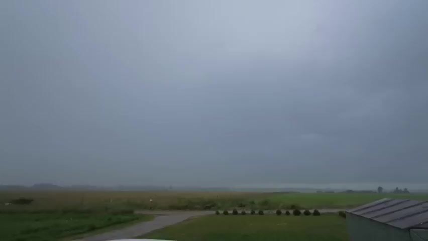 The video captures a wide shot of a rural landscape under a cloudy, overcast sky. The foreground shows a patch of green grass and the corner of a dark green shed on the right. A grey asphalt driveway leads away from the viewer into the midground. The midground consists of green fields and a strip of taller grass or vegetation. The background is a flat expanse of fields fading into the distance, obscured by a hazy atmosphere. The sky is a uniform, dull grey, suggesting an impending storm or recent rain. There are no people or obvious signs of activity.