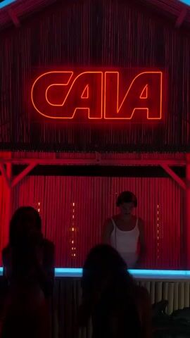 A vertical video captures a night scene in a club or bar, dominated by red and orange lighting. A large, bright orange neon sign reading "CALA" is mounted on a wooden structure at the top center, which also features a corrugated metal roof and red bamboo-like screens. Below the sign, a DJ is visible behind a counter with a blue light strip. The DJ, a person with short dark hair wearing a white tank top and headphones, is actively mixing music. In the foreground, two figures, silhouetted against the red light, are dancing, raising their arms and moving to the beat of the electronic music playing.