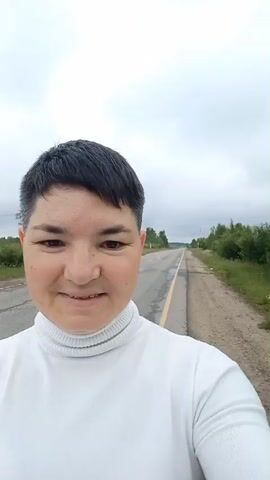 A woman with short dark hair and a white turtleneck is taking a selfie outdoors. She is standing on the side of a paved road that stretches into the distance under a cloudy sky. The road has a yellow line marking the lane. Green trees and foliage are visible on both sides of the road. The woman is smiling at the camera.
