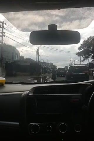 The video, shot from inside a vehicle, looks through the rearview mirror and windshield. The rearview mirror is in the upper left of the frame. Through the windshield, a street scene is visible with buildings, utility poles, and wires lining both sides of the road. Several motorcycles and cars are on the street, and people are walking around. The sky is cloudy. The interior of the car's dashboard is visible on the right side of the frame.