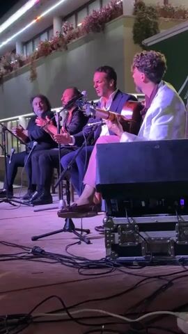 The video shows a group of four men performing music on a stage. From left to right, the first man is clapping his hands, the second man is also clapping his hands and singing into a microphone, the third man is playing a guitar and singing into a microphone, and the fourth man is playing a guitar. The men are dressed in formal attire. The stage is lit with purple and white lights. There are microphones, speaker equipment, and cables on the stage. The background shows a building with flowers on the balconies. The scene appears to be an outdoor performance at night.