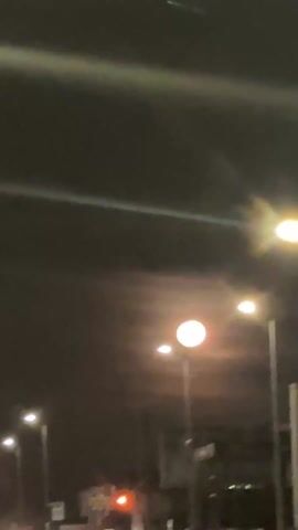 The video shows a full moon at night through the front windshield of a moving vehicle. The moon is bright white and surrounded by a soft orange glow. There are several streetlights visible in the foreground. The background is dark and unclear, but appears to be a street with buildings. The camera is shaky and the focus is blurred. The word "лисица" (lisitsa) is spoken, meaning "fox" in Russian. There is also music playing in the background.