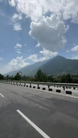 A POV shot captures a journey along a highway, curving gently to the left with majestic mountains in the distance. The sky is a vibrant blue, dotted with fluffy white clouds that cast shadows on the landscape. A white line marks the lane on the paved road, while a black and white striped guardrail lines the side of the highway. Trees and some buildings are visible in the valley below the mountains, and a yellow sign indicating a curve is posted on the side of the road. The sun is bright, suggesting it's daytime.