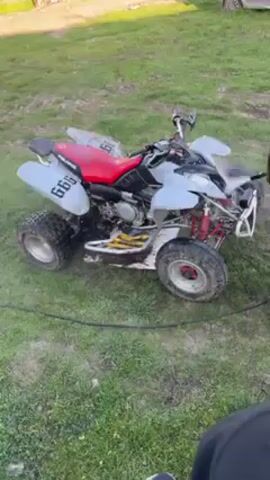 A man is pressure washing an ATV on a grassy surface. The ATV is white and gray with a red seat and the number "666" displayed on its side. The ATV appears muddy. A light gray car is parked in the background.