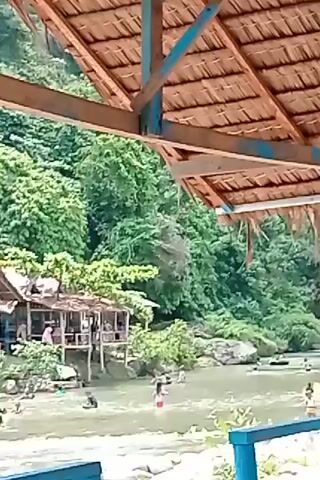 A video shows a river with people swimming and playing in the water. Several small huts with thatched roofs are located on the bank of the river, with some people sitting inside. The river is surrounded by lush green trees and vegetation, indicating a natural, possibly tropical, environment. The water appears shallow in some areas, allowing people to stand, while deeper sections are suitable for swimming and playing with inflatable tubes. There are rocks and stones visible in and around the river. The scene suggests a recreational area or resort where people are enjoying a day outdoors.