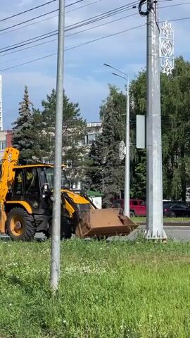 A yellow JCB backhoe loader is driving along a paved road, its front bucket lowered. The backhoe is in the foreground, with a grassy area and some construction debris to its left and in the foreground. Behind the backhoe, there are trees and buildings. A person is visible walking on the sidewalk in the background. The sky is clear and the sun is shining.
