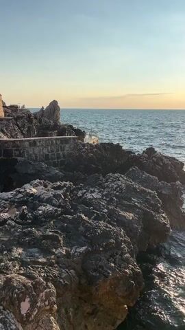 A video shows a rocky coastline with the ocean. The sun is shining and reflects on the water. There are some buoys in the water, one red and one white. The waves are gently lapping against the rocks. The sky is clear and blue. There are no people visible in the video.