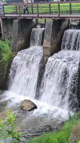 The video shows a multi-tiered waterfall with concrete structures and a wooden bridge overhead. Water cascades down from the top of the structure into a pool below. On the bridge, several people are visible, and there are cars parked and driving on a road behind the bridge. Green foliage and grass surround the waterfall and the area. The scene appears to be a public park or scenic spot.