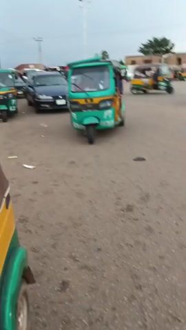 The video shows a turquoise and yellow auto-rickshaw driving down a dirt road. The auto-rickshaw is carrying passengers and luggage. The road is lined with trees and buildings. The sky is cloudy. There are other vehicles on the road, including cars and motorcycles. The auto-rickshaw appears to be in motion and is moving from left to right in the frame.
