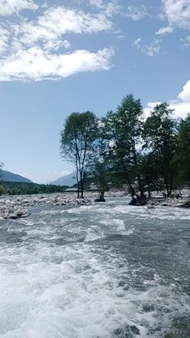 This media file depicts a natural scene with a river flowing through a rocky landscape. The water is white and foamy, suggesting it might be rapids or a waterfall. Along the riverbank, there are trees and rocks. In the background, a mountain range is visible under a bright, cloudy sky, indicating daytime.

A notable detail is a concrete pipe emerging from the bank and discharging water into the river. To the left of the pipe, there is a significant amount of trash and debris, including plastic bags and bottles, which are also scattered on the ground near the pipe and the riverbank. This contrast between the natural beauty of the river and mountains and the man-made pollution is a prominent element of the scene.

The overall impression is one of a potentially beautiful natural area marred by pollution. The clear sky and bright sunlight suggest pleasant weather. The presence of the pipe and the trash hints at human activity impacting the environment. There is no visible text in the media file. The setting appears to be a river in a mountainous region, with the location context suggesting Batahar, India.