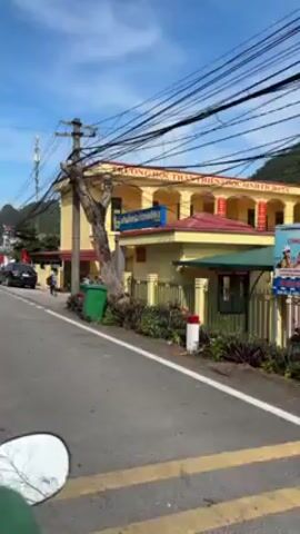 The video shows a street scene with a yellow building, which appears to be a school, based on the signage and the group of children wearing backpacks leaving the building's entrance. The building is surrounded by a fence, and there's a black car parked nearby. Power lines are heavily strung overhead.  The sky is partly cloudy. The camera is moving, likely from a vehicle passing by on the road.