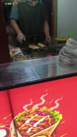 This is a close-up, slightly shaky, eye-level shot from behind a glass counter at a food stall. In the background, a person wearing gloves and a dark apron is cooking what appear to be rolled food items on a flat-top grill. Two rolled items are visible, one of which is being flipped with tongs. To the left of the grill are containers of ingredients, and to the right, a jar of sauce and a plastic bag. The foreground is dominated by a red surface with a stylized illustration of a rolled food item, possibly a wrap or burrito, topped with meat, lettuce, onions, and a drizzle of sauce, with steam or smoke depicted rising from it. The lighting suggests an indoor, possibly nighttime setting.