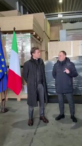 Two men stand in a warehouse setting with stacks of plywood and large packages in the background. The man on the left is wearing a long black coat over a gray suit and brown shoes. He appears to have a clerical collar. The man on the right is wearing a navy blue puffer jacket, gray pants, and black shoes.  An Italian flag and an EU flag are positioned to the left of the men. The man on the right is gesturing with his hand while speaking. The man on the left is listening attentively.