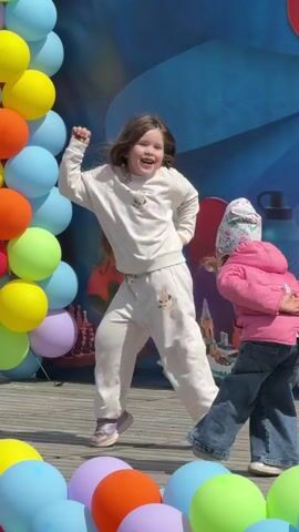 Two young girls are dancing on a wooden stage. The girl in the foreground is wearing a beige sweatsuit and sneakers, while the other girl has on a pink jacket, blue jeans, and a floral hat. Both are moving energetically, seemingly to music. Behind them is a large backdrop with musical instruments and a water bottle, framed by a balloon arch made of colorful balloons. The setting suggests an outdoor event or celebration.