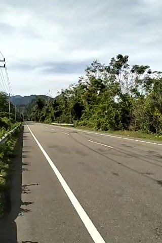 The video shows a two-lane asphalt road winding through a lush, green environment with a mountain range visible in the distance. The road has a solid white line marking the edge of the lane. A black SUV is traveling away from the camera down the road. On the left side of the road there is a metal guardrail and telephone poles. The sky is overcast with a mix of white and gray clouds.