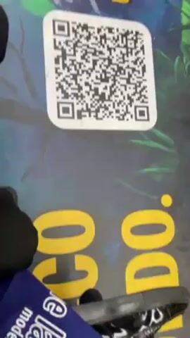 The video shows the interior of a moving subway train. The floor is covered with a colorful graphic design.  A large QR code is visible on the floor. Two pairs of feet, one wearing white sneakers and the other wearing black shoes, are standing near the QR code. The train appears to be carrying passengers. The text "INQUADRA E SBLOCCA UN RICORDO." is also printed on the floor near the QR code.  The overall environment suggests a promotional or interactive installation inside the train.