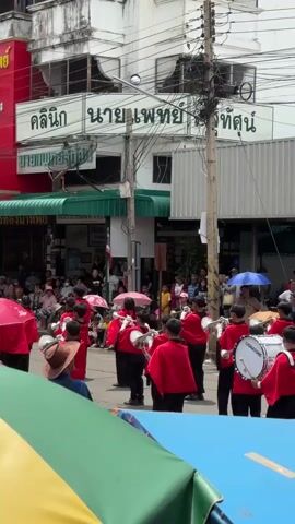This is a vertical, eye-level shot of a street parade featuring a marching band of children wearing red capes over black uniforms. The children are playing brass instruments, including tubas and trumpets, and snare drums. The drums are white with black logos that read "SIGNATURE". The band members are marching in formation on a paved street, with spectators on the left side of the frame watching the parade. The spectators are a mix of adults and children, some holding umbrellas. The buildings lining the street are two or three stories tall and appear to be shops or businesses. There are power lines and utility poles visible above the buildings. The sky is overcast. The foreground is occupied by colorful banners, one green and yellow, another blue, suggesting the viewpoint is from behind a barrier or a viewing area for the parade. The overall atmosphere appears festive and celebratory.