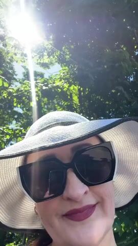 A woman wears a wide-brimmed white hat with a black band and large black sunglasses. She is outdoors with sunlight shining through green foliage from above. She is looking towards the camera and smiling faintly.