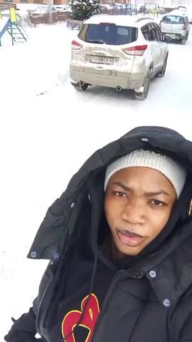 The video shows a person in a snowy environment. The person is wearing a black hooded jacket, a white knit hat, and headphones. They appear to be holding the camera and filming a selfie-style video, as their face is clearly visible in the foreground.  Snow covers the ground, and in the background, there are buildings, cars, and other people walking. The person is moving, and the camera angle shifts slightly, showing different parts of the snowy landscape.