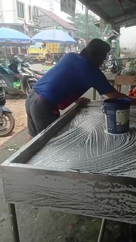 A person is cleaning a metal table with soap and water in an outdoor market. The person is wearing a blue t-shirt and dark pants and is using a pink sponge to scrub the table. The table is covered in soap suds. In the background, there are motorcycles, fruit and vegetables, and other market stalls. The person appears to be working diligently to clean the table.