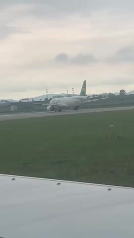 A passenger airplane with a white fuselage and green accents on its tail is visible on a tarmac. The plane is stationary, facing to the left. The foreground is dominated by a section of a white airplane wing, obscuring the bottom portion of the scene. The background features a grassy area and distant hills under a cloudy sky. The video is short, lasting only a few seconds, and appears to be filmed from inside another aircraft.