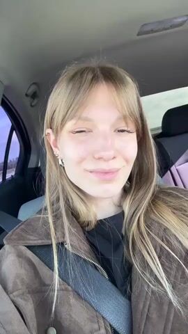 A young woman with long blonde hair and bangs is shown in a close-up selfie video, sitting in the driver's seat of a car. She is wearing a brown suede jacket, a black shirt, and a seatbelt. She smiles, briefly sticks out her tongue, and then smiles again. The car's interior and the outside scenery are visible in the background.