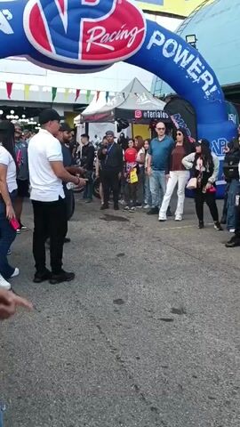 The video shows a street scene with a crowd of people gathered under a "VP Racing Powerkin" inflatable arch. Several individuals are walking around. A motorcycle carrying two people drives through the crowd. The location appears to be an outdoor event. The camera is handheld and moves with the person filming, showing various faces in the crowd. A person looks directly at the camera.