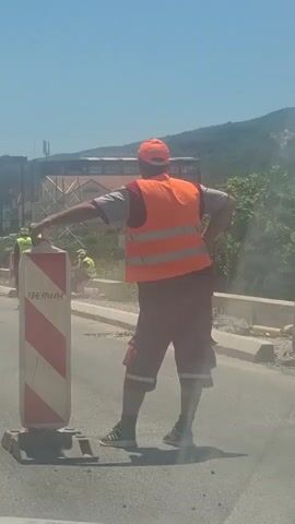 The video shows road construction in a town on a sunny day. A road worker wearing a bright orange safety vest and hat leans on a red and white striped road marker. In the background, a group of workers wearing yellow safety vests and hats are working on the side of the road. Some are using shovels, and one is using a saw. A large bus is driving in the background. Buildings and green hills can be seen in the distance. The air is dusty from the construction.