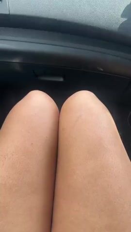 The video opens with a close-up view of a person's bare knees and thighs, showing visible body hair, from a first-person perspective inside a car. The camera then pans up to reveal the dashboard and the view through the front windshield. The car is moving on a two-lane road that curves through a lush green landscape. Trees line both sides of the road, and distant mountains are visible under a partly cloudy sky. Other vehicles can be seen ahead on the road. A smartphone, mounted on the right side of the dashboard, displays a navigation app with a visible estimated travel time of "2hr 37 min." The scene depicts a daytime road trip in a scenic, mountainous region near Mehadia, Romania.