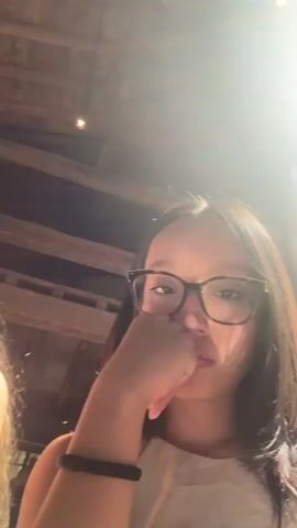 The media file contains a series of close-up selfies taken outdoors. The main subject is a young woman wearing glasses and a light-colored top, with her hand resting on her chin, suggesting a thoughtful or contemplative mood. She is accompanied by other individuals, though their faces are partially obscured or out of focus in some shots. One of these individuals, a young woman with blonde hair, is visible in the foreground of one selfie. Another shot includes a glimpse of a young man with curly hair in the background.

The setting appears to be a public space with rustic wooden beams visible in the background, possibly indicating an outdoor seating area at a restaurant or cafe. The bright sunlight streaming in suggests it is daytime, likely late morning or early afternoon. The weather appears to be clear and sunny.

There is no visible text in the media file. The overall impression is of a casual outing among friends on a bright, sunny day.