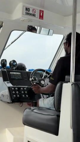 A man wearing sunglasses and a black t-shirt is steering a boat. He is seated in a black leather seat. The boat's interior is white with a dashboard of various controls. There are two small fans on the dashboard. A "NO SMOKING" sign is visible above the windshield. Outside the windshield, the sky is overcast.