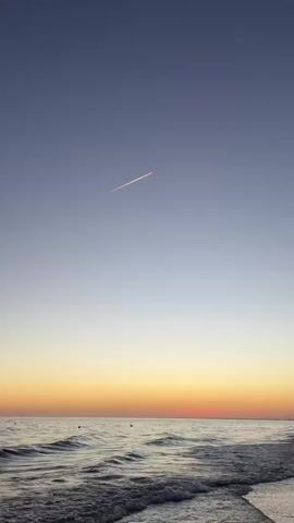 The video shows a serene beach scene at sunset. The sky transitions from a deep blue at the top to a gradient of pale yellow and orange near the horizon. A thin, white contrail from an airplane is visible stretching diagonally across the upper portion of the sky. The ocean is calm with gentle waves rolling towards a sandy shore. The water is a dark, reflective hue, mirroring the colors of the sunset. A few small dark shapes, possibly people or buoys, are scattered in the water. The overall atmosphere is peaceful and tranquil.