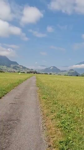 The video shows a paved path leading through a grassy field towards distant mountains under a bright blue sky with scattered white clouds. The path is in the foreground, with the tall green grass of the field to its right. In the middle ground, the path disappears into the field, and the landscape opens up to rolling hills and mountains. Some buildings and a construction crane are visible in the distance on the right side of the frame, near the base of the mountains. The overall impression is a serene, rural landscape on a sunny day.