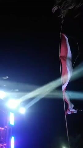 The video opens with a flag, possibly the Indonesian flag, waving in the foreground against a dark night sky. Several bright spotlights cut through the darkness, illuminating what appears to be a large sound system or stage setup. The lights are in motion, shifting colors from white and blue to green, yellow, pink, and purple. A crowd of people, many wearing t-shirts with a similar logo, are gathered in front of the sound system, some appearing to be dancing or observing. Trees and foliage frame the scene, suggesting an outdoor event. The camera pans and tilts, capturing the dynamic light show and the atmosphere of a nighttime gathering. The focus shifts to the people and the overall environment as the video progresses, with the lighting continuing to change and create a vibrant ambiance. Some individuals are seen interacting with the lights or each other, while others are simply present, enjoying the event. The video ends with the lights still actively changing colors, casting a colorful glow on the surrounding area and the attendees.