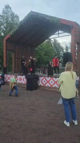 The video shows an outdoor concert stage decorated with greenery on its roof. On stage, a band is performing. A male guitarist stands to the left, a female singer is in the center, and another male is to the right. Behind them, a drummer is visible. The stage is adorned with a red and white patterned fabric along its front. In front of the stage, several children are dancing. To the left, a young boy wearing a colorful striped jacket is facing the stage. To the right, two girls in casual clothing are dancing. In the foreground, a woman with blonde hair wearing a yellow jacket and blue jeans is standing and watching the performance. The ground in front of the stage appears to be dirt. Trees are visible in the background, and the sky is overcast.